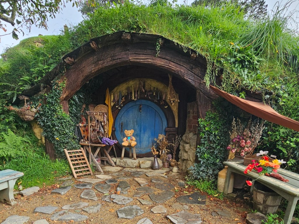 Patch Bear sitting outside a Hobbit home with a round blue door, flowers, tools, and a grass-covered roof.