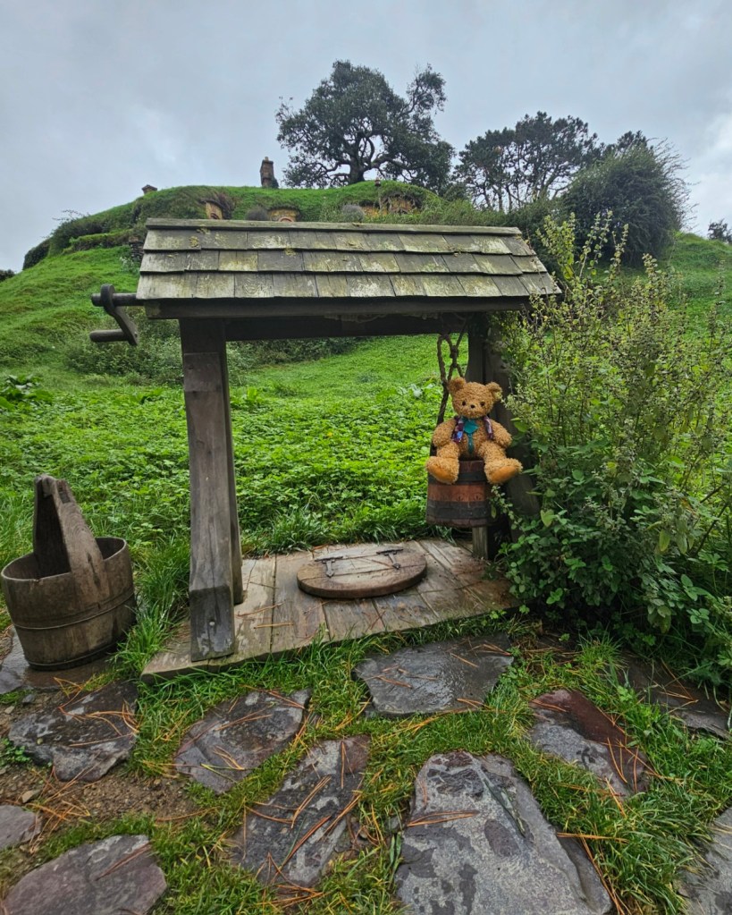 Patch sitting in the bucket, beside the closed well, with hobbit homes in the background.