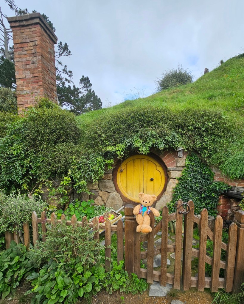 Patch sitting on the gate in front a of yellow Hobbit door, with a wheelbarrel of fruit and a chimney in the background.